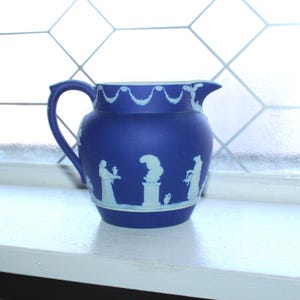 May include: A blue ceramic pitcher with white relief designs of figures and a floral pattern around the rim.