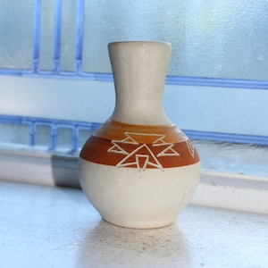 May include: A small, white ceramic vase with a brown band and geometric design. The vase has a narrow neck and a rounded body.
