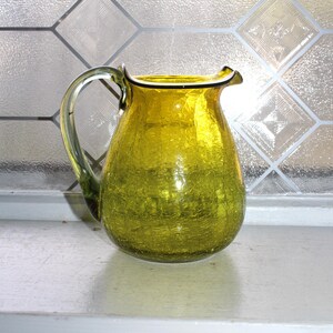May include: A yellow-green glass pitcher with a crackle finish. The pitcher has a curved handle and a slightly flared rim. The pitcher is approximately 8 inches tall and is sitting on a white surface.