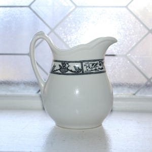 May include: A white ceramic pitcher with a curved handle and a spout. The pitcher has a decorative band of black and white floral and animal designs around the middle. The pitcher is sitting on a white surface.