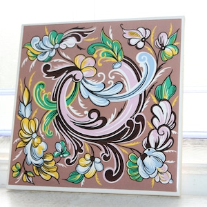 May include: A square ceramic tile with a brown background and a floral design in shades of pink, blue, green, and yellow. The design features stylized flowers and leaves in a swirling pattern.