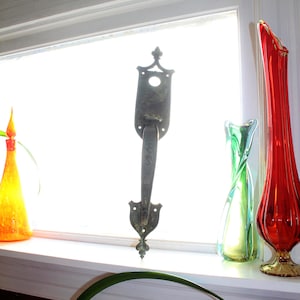 May include: A vintage metal door handle with a decorative design, displayed on a windowsill with three glass vases. The vases are in shades of green, red, and orange.