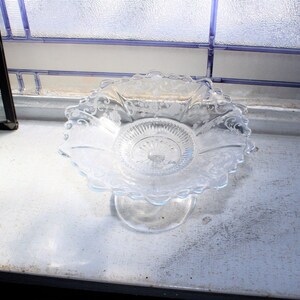 May include: A clear glass pedestal dish with a scalloped edge and intricate etched designs. The dish is on a white surface.