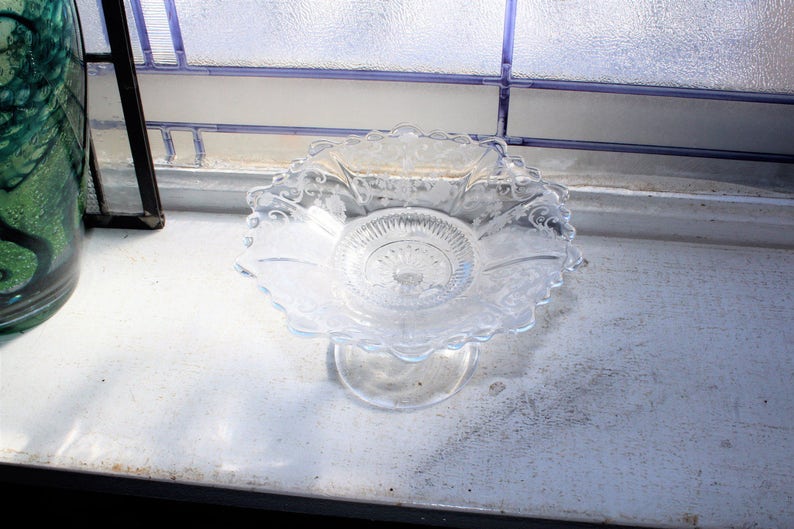May include: A clear glass pedestal dish with a scalloped edge and intricate etched designs. The dish is on a white surface.