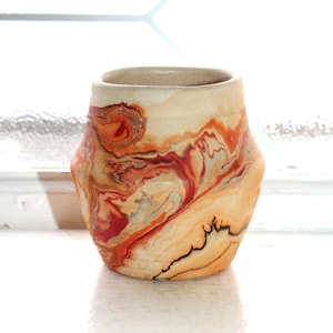 May include: A ceramic vase with a distinctive marbled pattern in orange, red, and cream hues. The vase has a hexagonal form and a neutral-toned rim. The interior is a light beige colour.