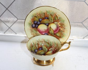 May include: A vintage teacup and saucer set with a fruit design. The cup and saucer are cream-colored with a gold trim. The fruit design includes peaches, grapes, and leaves. The cup sits on a small gold-colored stand.