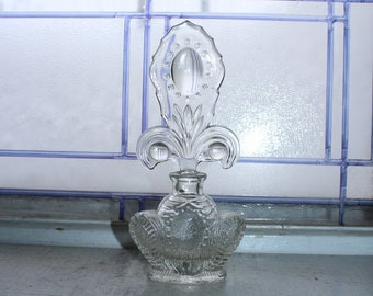 Può includere: Un flacone di profumo in vetro trasparente con un tappo decorativo a forma di giglio. La bottiglia presenta un design ornato in rilievo e poggia su una superficie azzurra. Lo sfondo presenta una finestra con finiture blu.