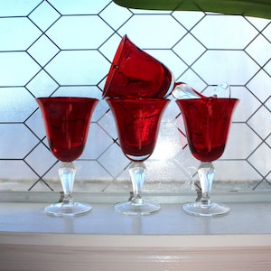 May include: Four red wine glasses with clear stems and bases are arranged on a white surface. The glasses have a tulip-shaped bowl. One glass is upside down, and another has a broken stem. The background features a window with a geometric pattern.