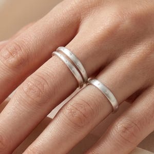 Può includere: Tre anelli color argento con finitura opaca e spazzolata. Due anelli sono sovrapposti sul dito medio, mentre un singolo anello abbinato è indossato sull'anulare. Ogni fascia presenta un design semplice e sottile con una superficie testurizzata.