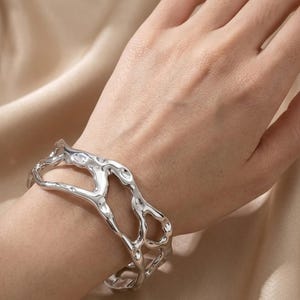 May include: A silver cuff bracelet with an abstract, open design. The bracelet features an uneven, textured finish and is worn on a wrist. The jewellery item is made of metal and has a contemporary, artistic style.
