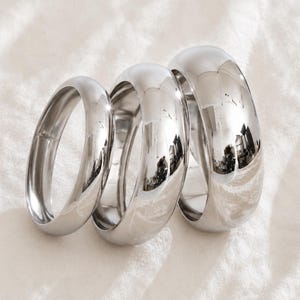 May include: Three polished silver-toned metal rings of varying widths arranged in a row. Each ring features a smooth, rounded, high-shine surface that reflects the surrounding light, creating a clean and reflective appearance across the set.