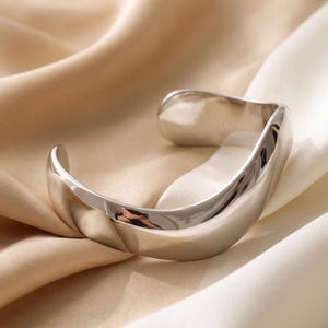 May include: A polished silver-tone cuff bracelet with a smooth, undulating wave-like shape. The metal surface is highly reflective, showing clear highlights and shadows that emphasise its curved, open-ended design and solid construction.