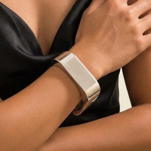 May include: A wide, polished silver-tone cuff bracelet with a smooth, reflective surface and a curved, rectangular profile. The bracelet features a solid, minimalist construction that wraps around the wrist, showcasing a clean, metallic finish.