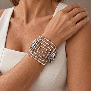 Può includere: Un ampio bracciale rigido color argento caratterizzato da un prominente design a spirale quadrata. La superficie metallica presenta una finitura martellata e testurizzata su tutto il motivo geometrico, con spirali più piccole sui lati.