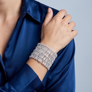 Può includere: Un ampio bracciale a fascia color argento caratterizzato da un intricato design in metallo intrecciato traforato. Il bracciale presenta una struttura flessibile simile a una rete, composta da numerosi piccoli anelli metallici interconnessi.