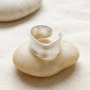 Può includere: Un anello color argento con una fascia ampia e curva. L'anello è appoggiato su una pietra liscia e chiara. L'anello ha il numero "925" stampato all'interno.