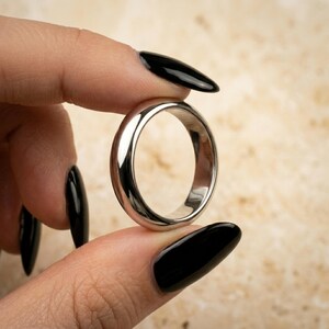 Könnte beinhalten: Ein polierter silberfarbener Metallring mit einem glatten, abgerundeten Band und einer hochglänzenden, reflektierenden Oberfläche. Der Ring zeichnet sich durch eine einfache, solide Konstruktion mit gleichmäßiger Breite über das gesamte kreisförmige Band aus.