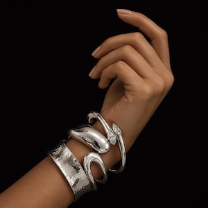 Pode incluir: Um conjunto de três pulseiras prateadas no pulso. Uma é um bracelete largo com textura martelada, outra é uma pulseira rígida espessa e lisa de forma orgânica, e a terceira é uma pulseira aberta fina com duas pedras transparentes em lapidação marquise.