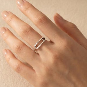 Può includere: Un anello color argento a forma di spilla da balia. L'anello è indossato su un dito, mostrando il suo design minimalista. Il metallo dell'anello ha una finitura lucida, che riflette la luce. L'anello è un gioiello.