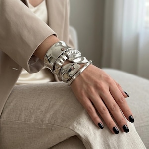 Pode incluir: Uma coleção de pulseiras de metal prateado com vários designs adorna um pulso. As pulseiras têm uma superfície polida e refletora. Os designs incluem uma faixa larga e lisa, um padrão semelhante a uma corrente e elementos orgânicos em forma de folha. As unhas estão pintadas de preto.