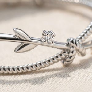 Può includere: Un anello color argento caratterizzato da un delicato motivo a ramo con due foglie lisce e una piccola pietra preziosa rotonda e trasparente incastonata a quattro griffe, abbinato a una fascia con texture a corda intrecciata.