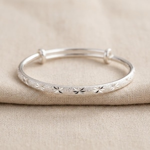 Puede incluir: Un brazalete ajustable de tono plateado con una superficie texturizada y esmerilada, decorado con un patrón repetitivo de estrellas de ocho puntas grabadas. El brazalete cuenta con dos bucles deslizantes superiores para ajustar.