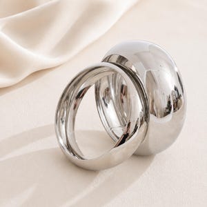 Pode incluir: Dois anéis de metal prateado polido com uma superfície lisa e reflexiva. Os anéis são circulares com um design de banda grossa e arredondada. O metal tem um acabamento de alto brilho que cria reflexos claros do ambiente ao redor.