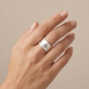 May include: A wide, silver-coloured band ring with a smooth, matte finish and a slightly concave, curved surface. The ring features a continuous, solid metal construction that wraps around the finger, showcasing a clean, polished look without any stones.