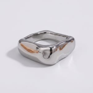 Può includere: Un anello color argento con una fascia di forma quadrata caratterizzata da una superficie liscia, lucida e irregolare che ricorda un liquido. Il metallo riflette la luce con una finitura brillante, creando riflessi morbidi sui bordi.