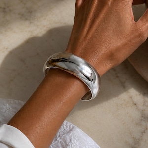 Könnte beinhalten: Ein breites, poliertes Silberarmband. Das Armband hat eine glatte, reflektierende Oberfläche und eine abgerundete Form. Licht reflektiert sich auf der Oberfläche und erzeugt Glanzlichter und Schatten. Das Armband wird am Handgelenk getragen.
