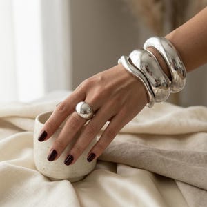 Può includere: Una mano ornata da un grande anello d'argento e tre braccialetti spessi abbinati. I gioielli sono molto lucidi e riflettenti. La mano poggia vicino a una piccola tazza di ceramica bianco sporco su un tessuto color crema.