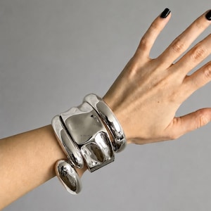 Può includere: Un set di braccialetti e un anello in metallo argentato. I braccialetti hanno un design scultoreo e massiccio con elementi arrotondati e rettangolari. L'anello è di forma ovale. I gioielli sono indossati su un polso su sfondo grigio.