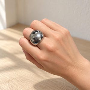 Könnte beinhalten: Ein klobiger, silberfarbener Ring mit einer glatten, abgerundeten und bauchigen Oberfläche. Das Metall hat ein hochglänzendes Finish, das Licht und umgebende Formen reflektiert und so ein markantes, massives und nahtloses Erscheinungsbild erzeugt.