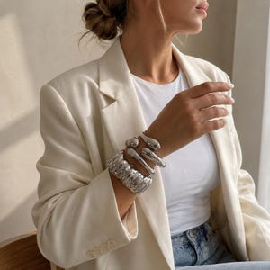 Può includere: Bracciali color argento in vari design adornano un polso. I bracciali includono un polsino largo e strutturato e diversi braccialetti lisci a forma organica. La persona indossa un blazer e una maglietta bianchi.