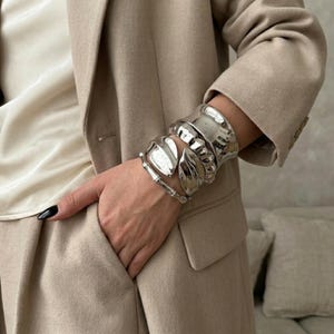 Può includere: Una collezione di braccialetti in metallo argentato. I braccialetti hanno vari design, tra cui un polsino largo, forme organiche e un braccialetto in stile bambù. La persona indossa un blazer beige e un top color crema.