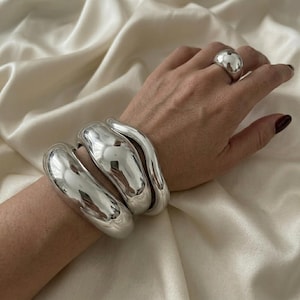 Può includere: Un set di due bracciali rigidi spessi in metallo color argento lucido con una forma organica liscia e arrotondata indossati al polso, accompagnati da un anello abbinato in metallo color argento spesso e arrotondato con finitura riflettente.