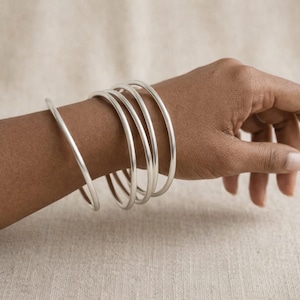 Può includere: Un set di quattro bracciali rigidi color argento, lisci e lucidi. Ogni bracciale presenta un design circolare semplice e arrotondato, con uno spessore costante e una finitura metallica riflettente, creando un aspetto sovrapposto.