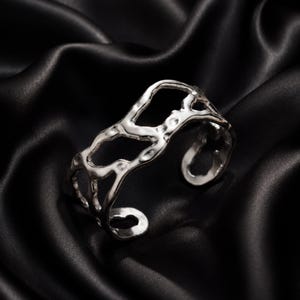Può includere: Un bracciale a polsino color argento con un design aperto e organico. Il bracciale ha una superficie irregolare e strutturata, su uno sfondo nero e setoso. Il gioiello è un accessorio di tendenza.
