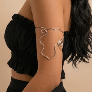 Può includere: Bracciale argentato con un design astratto e fluido. Il bracciale avvolge la parte superiore del braccio, con una serie di linee e spirali interconnesse. Il gioiello è indossato su uno sfondo scuro.