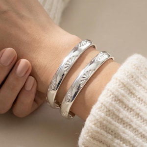 Può includere: Due braccialetti a polsino argentati incisi con un motivo a raggiera. I braccialetti sono indossati su un polso, con un maglione color crema sullo sfondo. I braccialetti sono realizzati in metallo lucido. I braccialetti hanno un diametro di circa 6 cm.