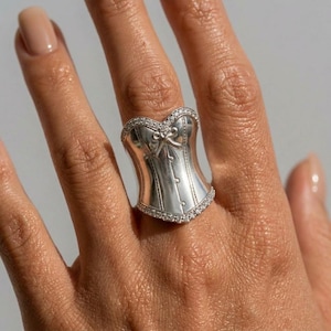 Può includere: Anello a corsetto argentato con un design dettagliato. L'anello presenta una forma a corsetto con un fiocco e piccoli abbellimenti rotondi. I bordi dell'anello sono bordati da piccole pietre trasparenti. L'anello è indossato su un dito.