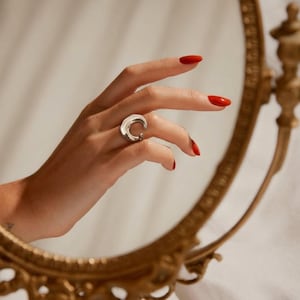 Può includere: Un anello argentato con un design curvo e aperto è indossato su una mano con smalto rosso. L'anello è il punto focale, che riflette la luce. Lo sfondo include uno specchio con cornice dorata.