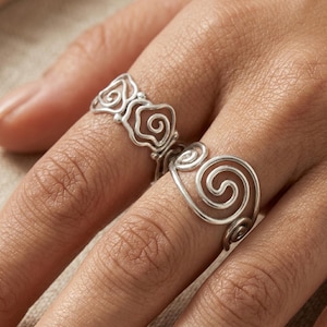 Può includere: Due anelli in argento indossati su dita adiacenti. L'anello superiore presenta un motivo ripetitivo di piccole spirali in filo metallico aperto. L'anello inferiore mostra un grande design a spirale realizzato in filo d'argento lucido.