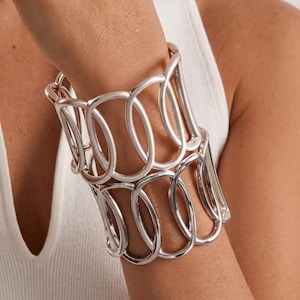 Può includere: Un ampio bracciale a fascia caratterizzato da un motivo traforato ripetitivo di anelli ovali intrecciati color argento. Il design è composto da due file sovrapposte di queste forme metalliche lisce e arrotondate che creano una struttura.