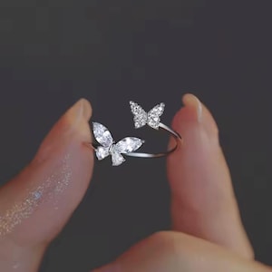 Könnte beinhalten: Ein silberner Ring mit zwei Schmetterlingsmotiven. Ein Schmetterling hat ein birnenförmiges Steindesign, der andere ist kleiner und strukturiert. Der Ring wird zwischen den Fingern vor einem dunklen Hintergrund gehalten. Ein zarter Schmuck.