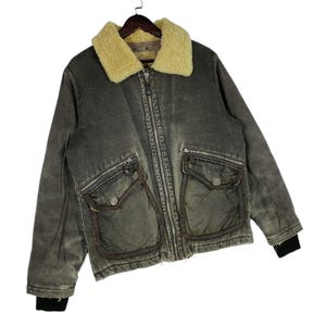 May include: An olive-green vintage jacket with a cream shearling collar. The jacket has a full zip, multiple pockets, and black ribbed cuffs. The jacket has a worn, distressed appearance, giving it a retro feel.