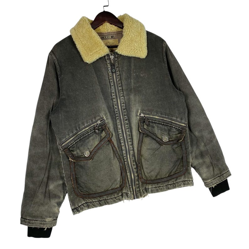 May include: An olive-green vintage jacket with a cream shearling collar. The jacket has a full zip, multiple pockets, and black ribbed cuffs. The jacket has a worn, distressed appearance, giving it a retro feel.