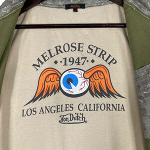 Puede incluir: Camiseta beige con el texto "MELROSE STRIP 1947" sobre un dise&ntilde;o de ojo con alas naranjas. El texto "LOS ANGELES CALIFORNIA" y el logotipo de Von Dutch est&aacute;n debajo. La camiseta est&aacute; dentro de una chaqueta verde y gris.