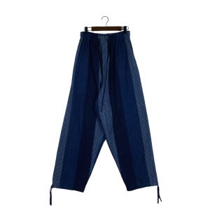 May include: Loose-fitting trousers in shades of blue featuring a vertical striped pattern with alternating solid dark blue panels and lighter blue textured panels. The pants have an elastic waistband with a drawstring and adjustable ties at the hems.