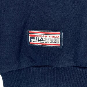 May include: A dark navy blue fabric garment featuring a rectangular patch with red and white horizontal stripes. The patch displays the text BIELLA ITALIA, FILA SPORT, and MODA NELLA VITA SPORTIVA in black and red lettering on a white background.